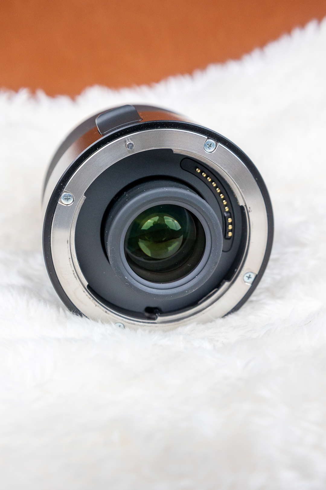 Rear view of a camera lens showing the metal mount, electronic contacts on the right, and the glass element at center, resting on a white fluffy surface.