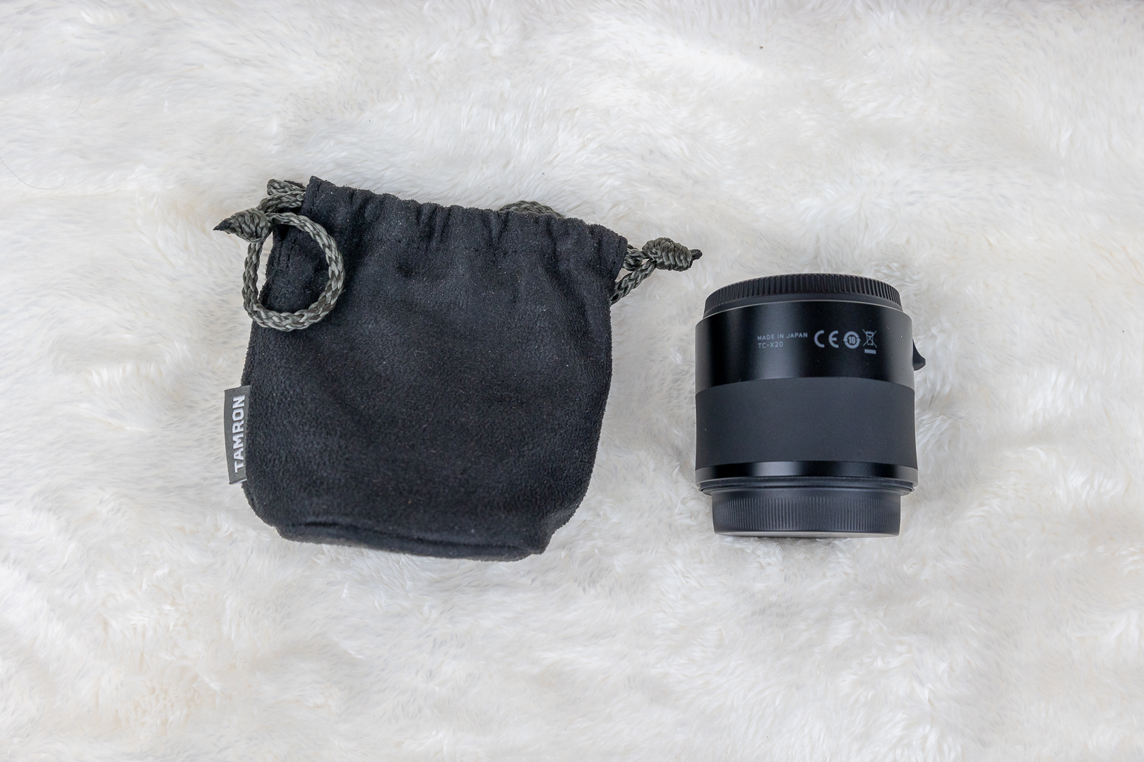 Tamron camera lens beside a black drawstring pouch on a white fluffy surface.