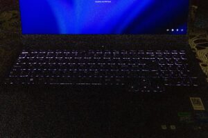 Close-up of a laptop keyboard with purple backlight and a bright blue screen above it in a dim room.